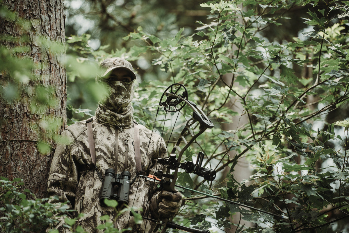 Bowhunter in S-18 Camo standing against a pine trunk in green timber, compound bow raised, early season.