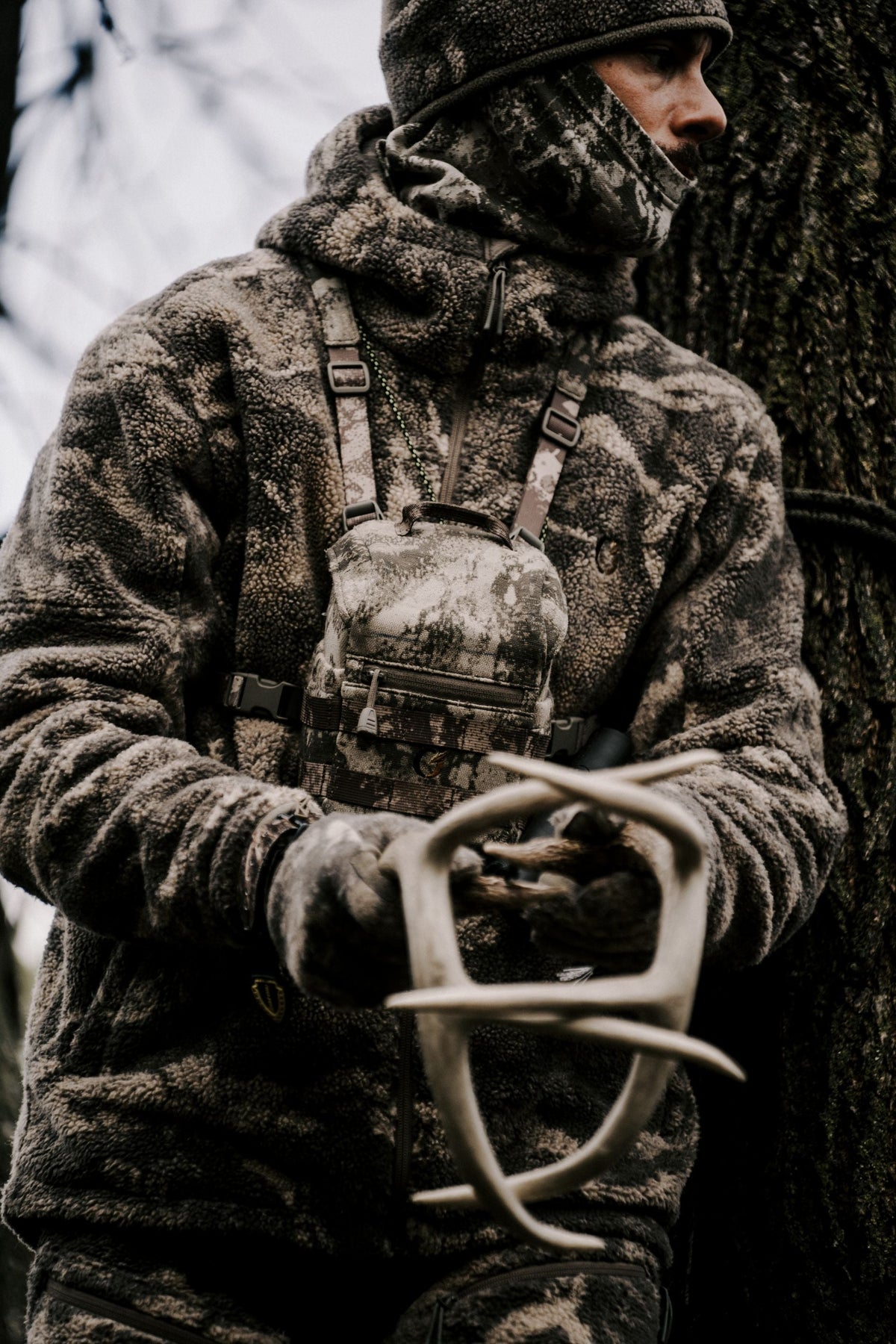 Hunter in S-18 Camo wearing the Silent Binocular Harness System, holding a shed antler by a tree