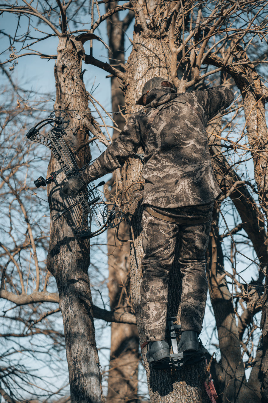 Bowhunter in S-18 Camo on a VantageTech stand platform in a bare hardwood, compound bow in hand