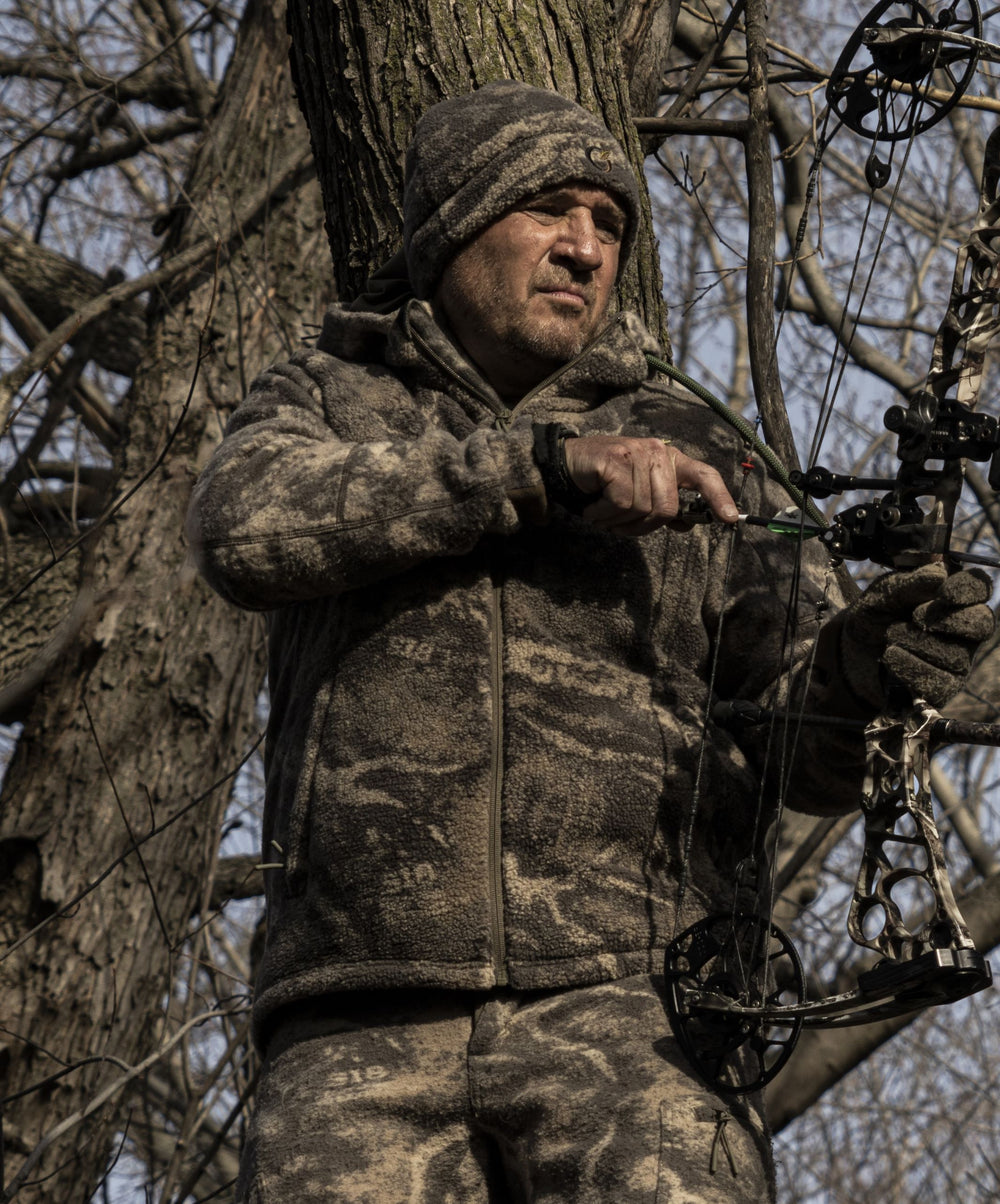 Bowhunter at full draw from a treestand in bare hardwoods wearing Zone7 Versa fleece.